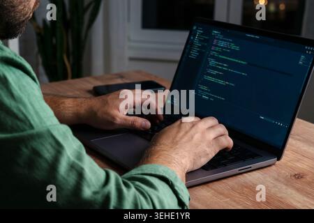 Man types on laptop in dimly lit room while receiving scam message on phone Stock Photo