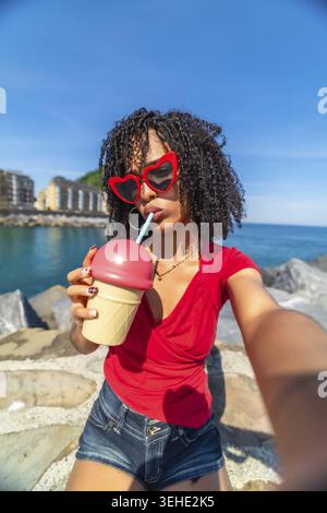 Young woman with heart shaped sunglasses and curly hair drinking a refreshing beverage from an ice cream shaped cup while taking a selfie during her s Stock Photo