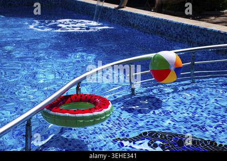 A colorful ball and an inflatable circle, with a watermelon pattern floating. in a sunny outdoor pool on a summer day, there are no people Stock Photo
