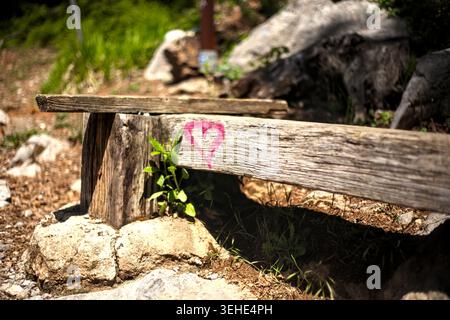 A small pink heart symbol painted on a weathered rustic wooden bench along a scenic mountain hiking trail. Stock Photo