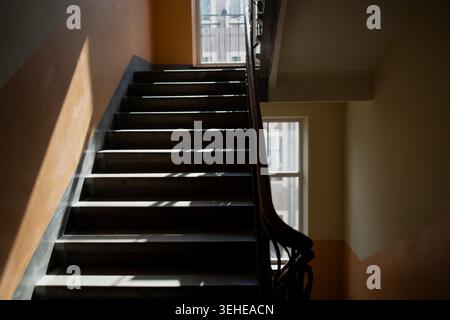 Staircase in old residential building with vintage wooden handrail and worn concrete steps leading upward to a glass window with dark metal security b Stock Photo