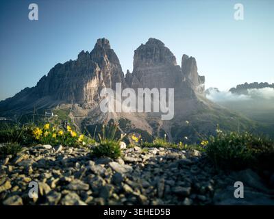 Sunlit view of the Tre Cime peaks rising above rocky slopes and alpine meadows, with soft morning light and mist creating a serene mountain scene. Stock Photo