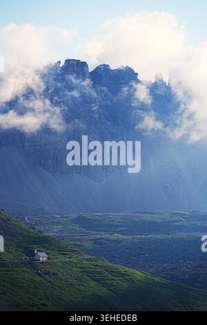 Soft morning mist drifts around dramatic rocky peaks above a green alpine valley, with a small mountain chapel standing quietly in the landscape. Stock Photo