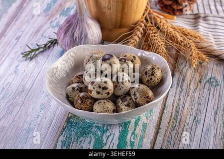 Still life of fresh speckled quail eggs in a white ceramic bowl. Rustic composition with garlic, wheat, and rosemary on a distressed wooden table. Nat Stock Photo