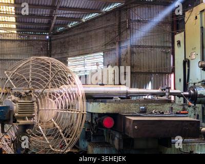 A large industrial lathe machine machining a heavy metal shaft inside an old factory workshop, with a big industrial fan in the foreground and light r Stock Photo
