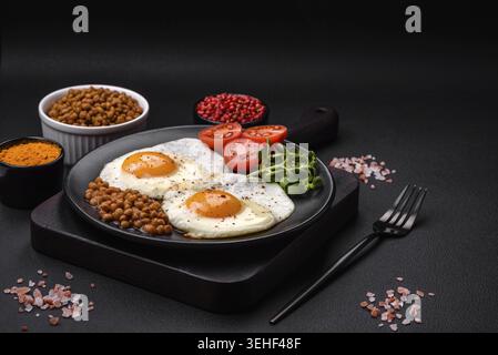 Food, Delicious hearty breakfast consisting of two fried eggs, canned lentils, microgreens with spices and herbs on a dark concrete background Stock Photo