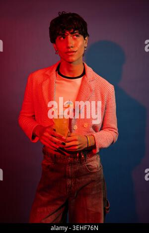 Young non-binary person holding a fun cocktail, celebrating personal style and identity in a vibrant setting Stock Photo