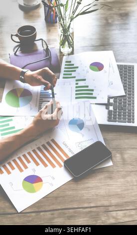 Person analyzes financial charts with focused attention. Hands hold pen near colorful pie and bar graphs. Laptop and smartphone lie nearby on wooden d Stock Photo