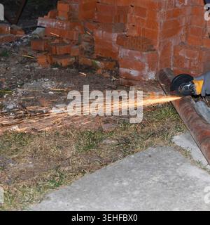 A person using a power tool to cut a metal pipe near a brick wall. Sparks are flying from cutting process. Scene is set outdoors on a concrete surface Stock Photo