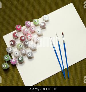 Art supplies arranged on a textured green background. Includes paint pots in various colors, three paintbrushes, and a blank canvas Stock Photo