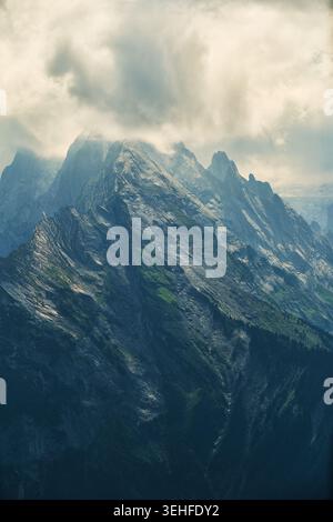 Dramatic alpine mountain peaks rise through clouds and mist above steep, rugged rocky ridges and forested slopes. Stock Photo