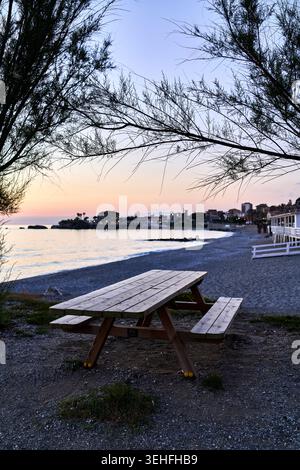 Wooden picnic table resting on a gravel beach, looking out at the calm sea during a colorful sunset or dusk Stock Photo