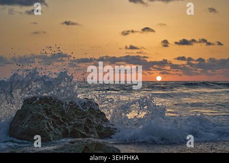 Ocean wave splashing over coastal rocks on a tranquil beach during a beautiful orange sunset, creating dynamic water spray Stock Photo