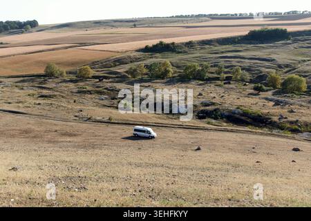 A white van parked on a dry, grassy landscape with rolling hills and sparse trees under a clear blue sky Stock Photo
