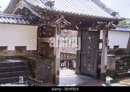 A traditional Japanese wooden gate with tiled roof covered in light snow, standing open to reveal a courtyard beyond. Snowflakes fall gently through Stock Photo