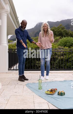 Mature man and senior woman standing on tiled patio gesturing to picnic blanket with wine glasses Stock Photo