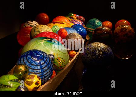 A collection of colorful handcrafted glass spheres displayed in wooden boat-like containers against a dark background. The spheres feature diverse Stock Photo