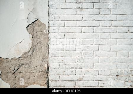 Weathered exterior wall divided between exposed worn plaster and white brick masonry, showing cracks, erosion and aged urban texture and background Stock Photo