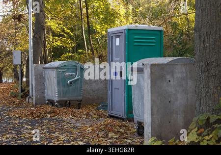 A portable toilet stands beside two waste bins in a wooded area. Autumn leaves cover ground, creating a natural setting for waste disposal Stock Photo