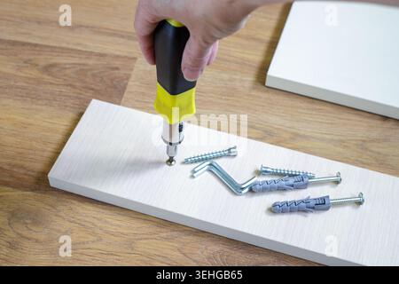 Hand screws a self-tapping screw into a furniture panel on a laminate floor. Fasteners, dowels and hex keys are ready for DIY home repairs and carpent Stock Photo