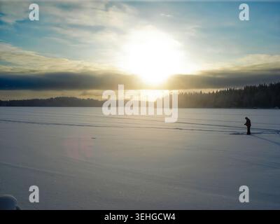A lonely fisherman on a snowy Finnish lake. The bright winter sun comes out from behind the clouds, illuminating the forest and the white surface of t Stock Photo