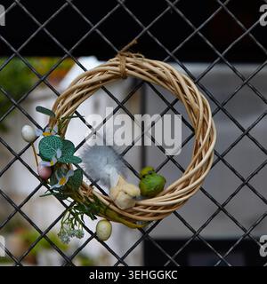 Easter wreath featuring vibrant eggs and a small bird, suspended on a black metal fence, surrounded by lush greenery in a garden environment Stock Photo