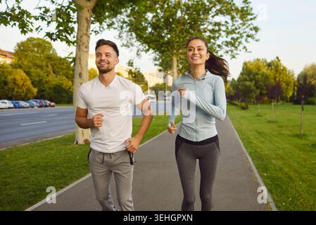 Smiling young couple jogging together along park path near city road, enjoying outdoor fitness run. Stock Photo