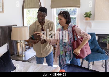 Diverse couple standing in bedroom examining passports and packing open suitcase with red handbag Stock Photo