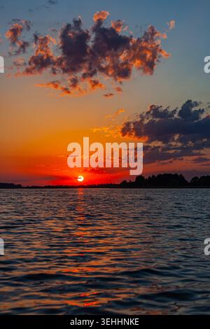 Fiery sunset above a calm lake with glowing reflections on rippling water, forest silhouettes and dramatic evening clouds. Stock Photo