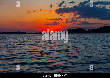 Golden sunset over a tranquil lake with glowing reflections, rippling water and dark forest silhouettes beneath dramatic clouds. Stock Photo