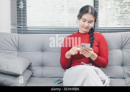 Young Asian woman using smartphone for online shopping at home, browsing ecommerce app on sofa with credit card nearby, mobile payment, cashless lifes Stock Photo