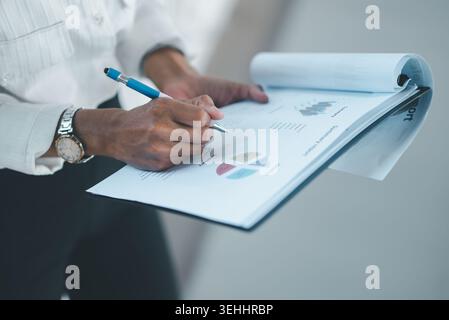 Close-up of hands writing notes on a business summary report with charts and graphs on a clipboard, highlighting data analysis, document review, finan Stock Photo