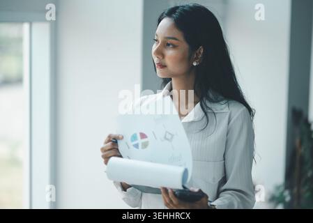 Professional woman holding a smartphone and report pages near a bright office window, representing business planning, mobile coordination, document re Stock Photo