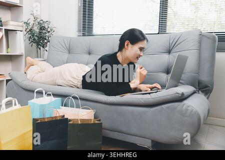 Young Asian woman lying on sofa using laptop for online shopping at home with colorful shopping bags, browsing ecommerce deals, digital retail lifesty Stock Photo