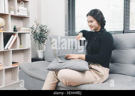 Young Asian woman using laptop and drinking coffee on sofa at home, remote work and online lifestyle concept, casual home office, freelance productivi Stock Photo