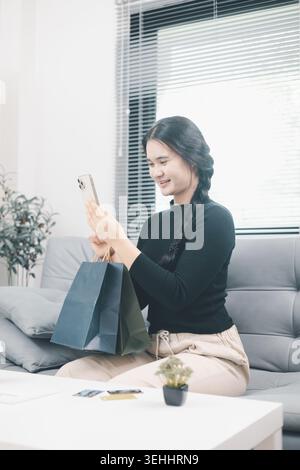 Young Asian woman holding shopping bags and using smartphone at home after online shopping, checking mobile app or order update, ecommerce retail life Stock Photo