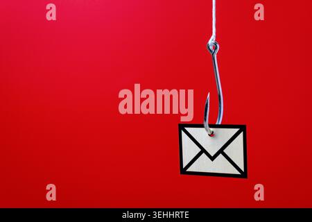 A metallic fishhook and envelope icon against a red background with copy space. Email phishing scams, cyber fraud, spam messages, and digital security Stock Photo