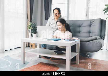 Two women work on a laptop at home while reviewing notes together, illustrating online guidance, remote learning, teamwork and modern home workspace s Stock Photo