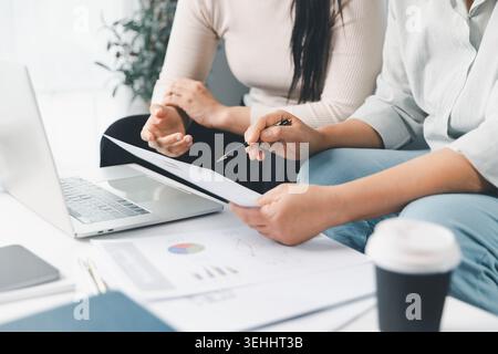 Two women review financial documents and charts beside a laptop at home, discussing budget planning, online consultation, business analysis, and house Stock Photo