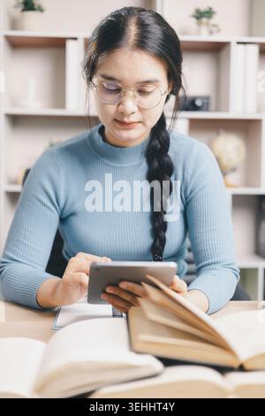 Young Asian woman in glasses using tablet while studying with open books at home desk. Digital learning, online research, self study, academic prepara Stock Photo