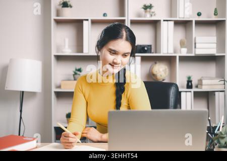 Young Asian woman writing notes while using laptop at home office desk. Smiling student in online learning, remote study, digital education, productiv Stock Photo