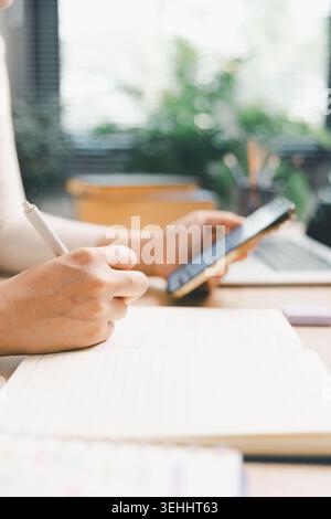 Close up of young Asian woman writing notes in notebook while using smartphone beside laptop at home desk. Mobile learning, online study, digital educ Stock Photo