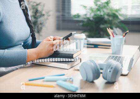 Young woman using smartphone over books beside laptop and headphones at home desk. Mobile learning, online study, digital research, multitasking, educ Stock Photo