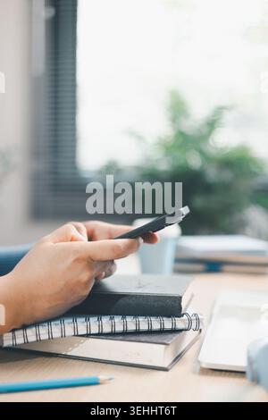 Young woman using smartphone over books beside laptop and headphones at home desk. Mobile learning, online study, digital research, multitasking, educ Stock Photo