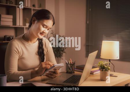 Young Asian woman writing notes beside laptop in cozy home office at night, showing remote work, online study, planning, productivity, concentration a Stock Photo