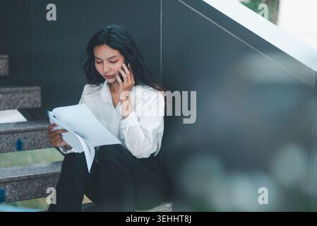 Businesswoman talking on a smartphone while reading reports on office stairs, showing mobile communication, multitasking, document review, problem sol Stock Photo