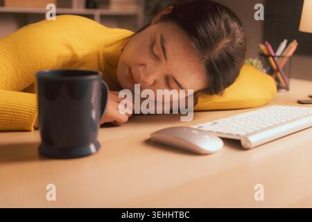Exhausted young Asian woman sleeping at desk beside keyboard and coffee in cozy home office at night, illustrating burnout, overtime, work fatigue, de Stock Photo
