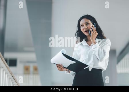 Professional woman speaking on a smartphone while reviewing documents on a clipboard in a bright office corridor, ideal for business communication, cl Stock Photo