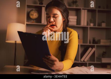 Sleepy young Asian woman yawning while reviewing clipboard documents in cozy home office at night, illustrating paperwork fatigue, overtime stress, de Stock Photo