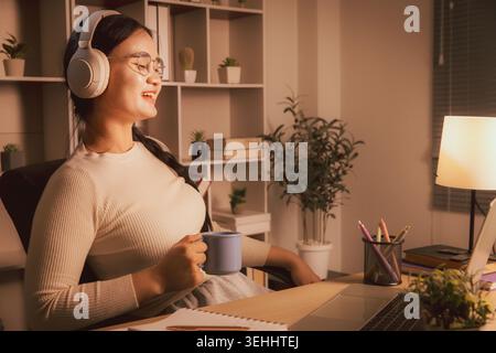 Smiling young Asian woman wearing headphones and holding coffee while relaxing by laptop in cozy home office at night, representing remote work break, Stock Photo
